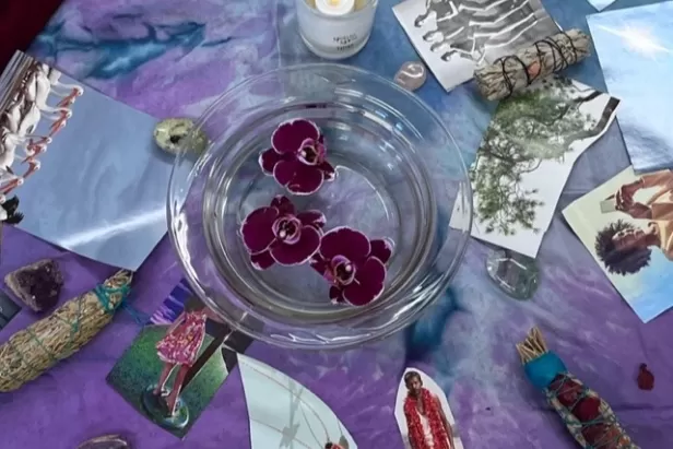 purple blanket with a bowl of water and red flowers floating, a candle, sage sticks and various pictures surrounding 
